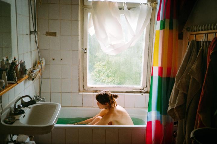 A woman sits in a bathtub with green water next to a window with a colorful shower curtain.