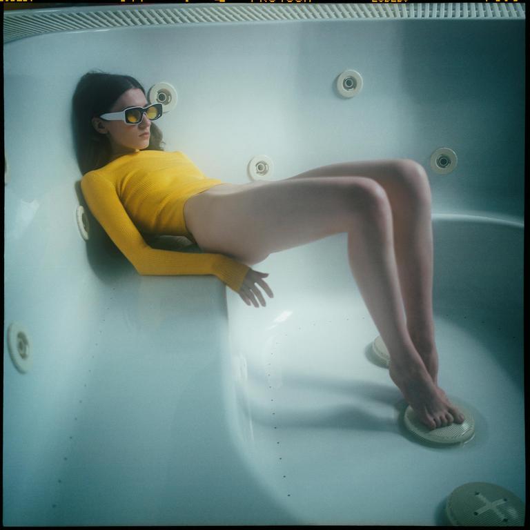 A woman wearing a yellow top and sunglasses is lying in a bathtub.