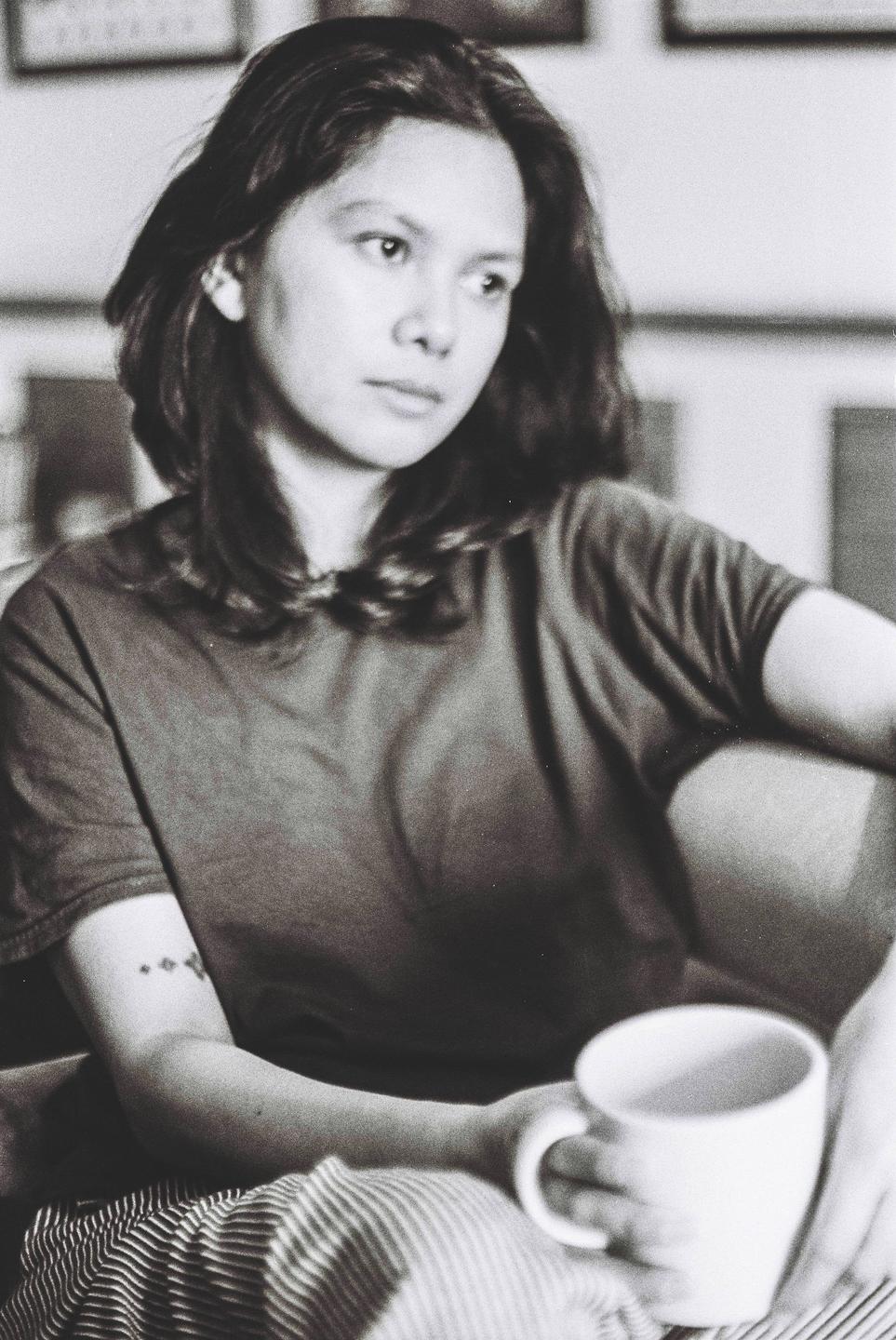 A woman with long hair sits holding a mug, looking away from the camera.