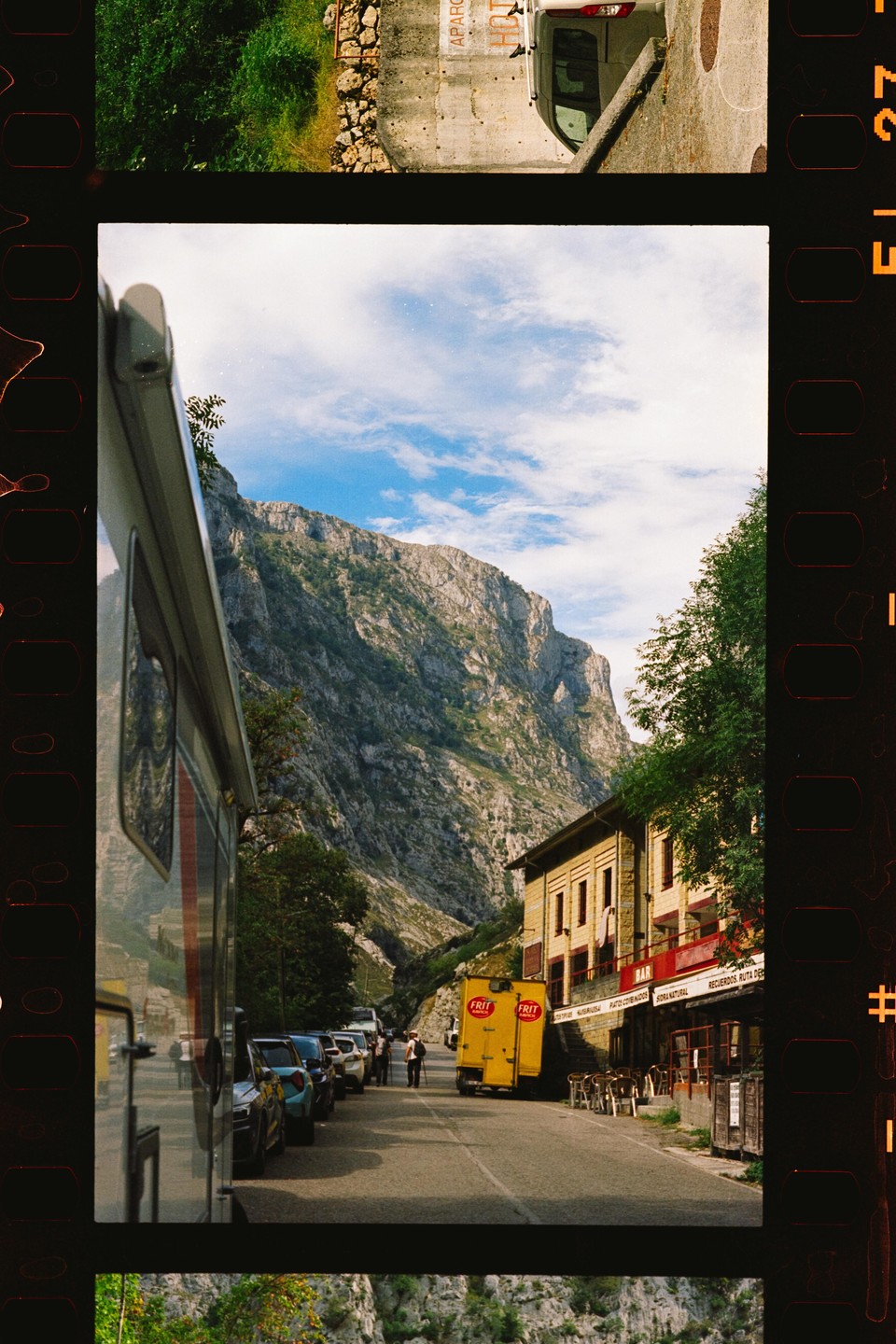 A street with parked cars and a yellow truck is nestled at the base of a large mountain, with a building and trees on the right.
