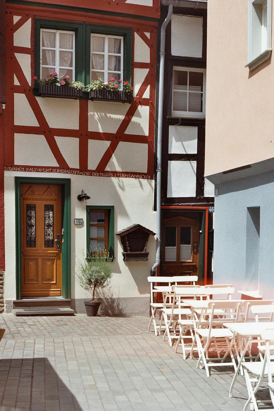 A half-timbered building with flower boxes and a wooden door stands next to a cafe with white tables and chairs on a paved street.