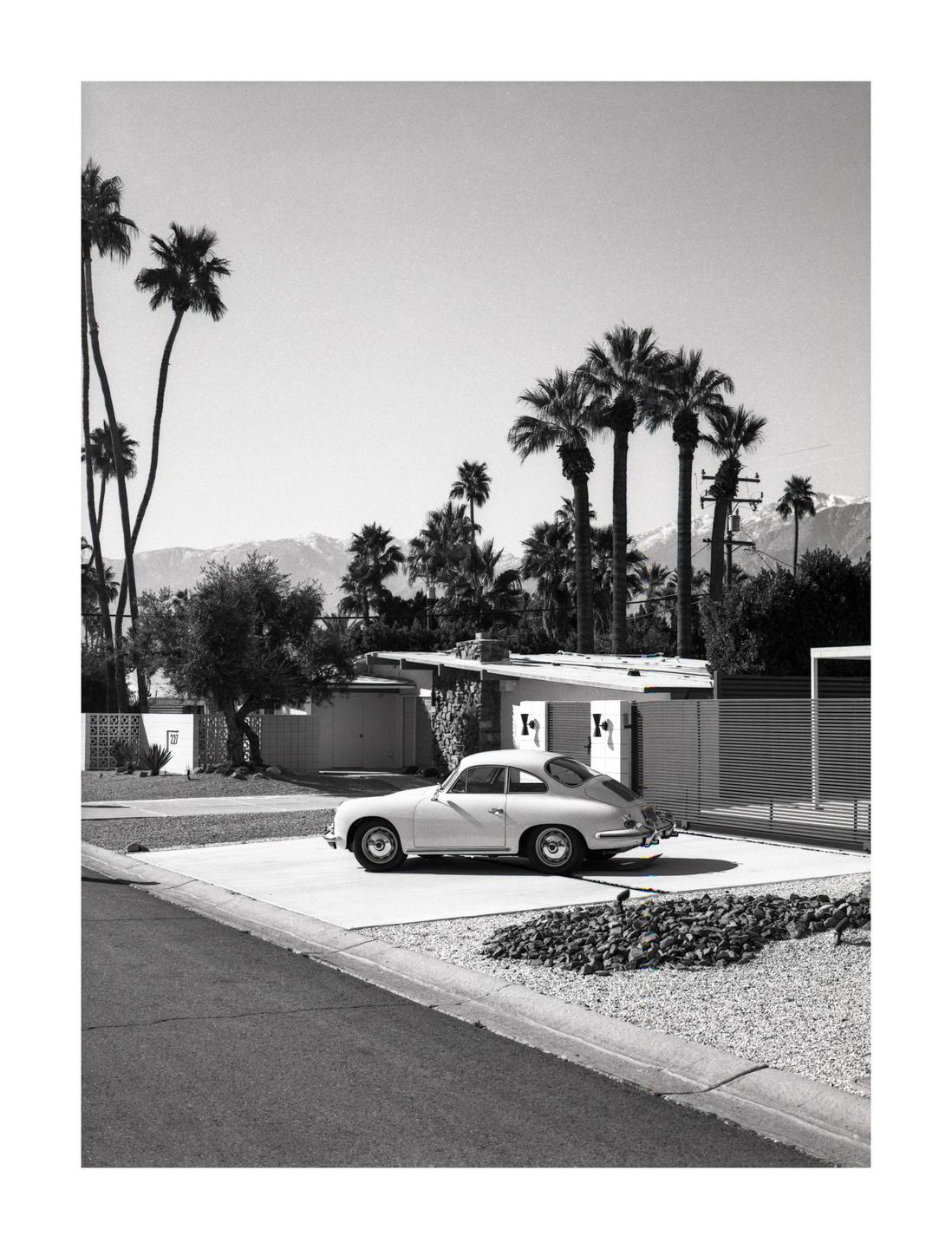 A vintage car is parked in a driveway in front of a house with palm trees and mountains in the background.