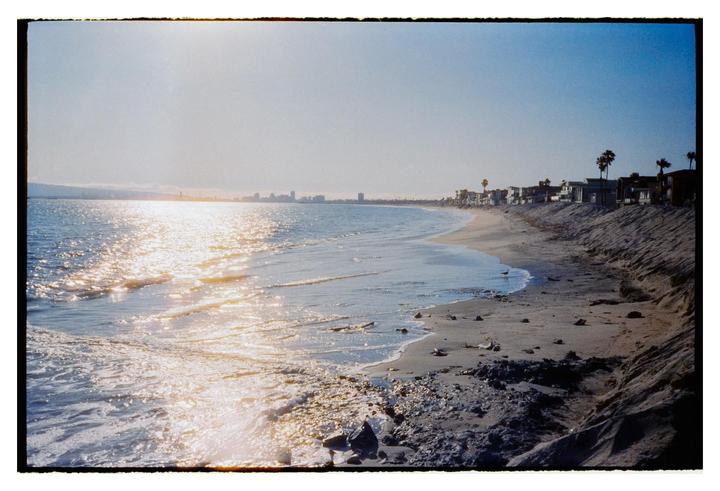 The sun reflects brightly on the ocean waves along a sandy beach with houses and a city skyline in the distance.