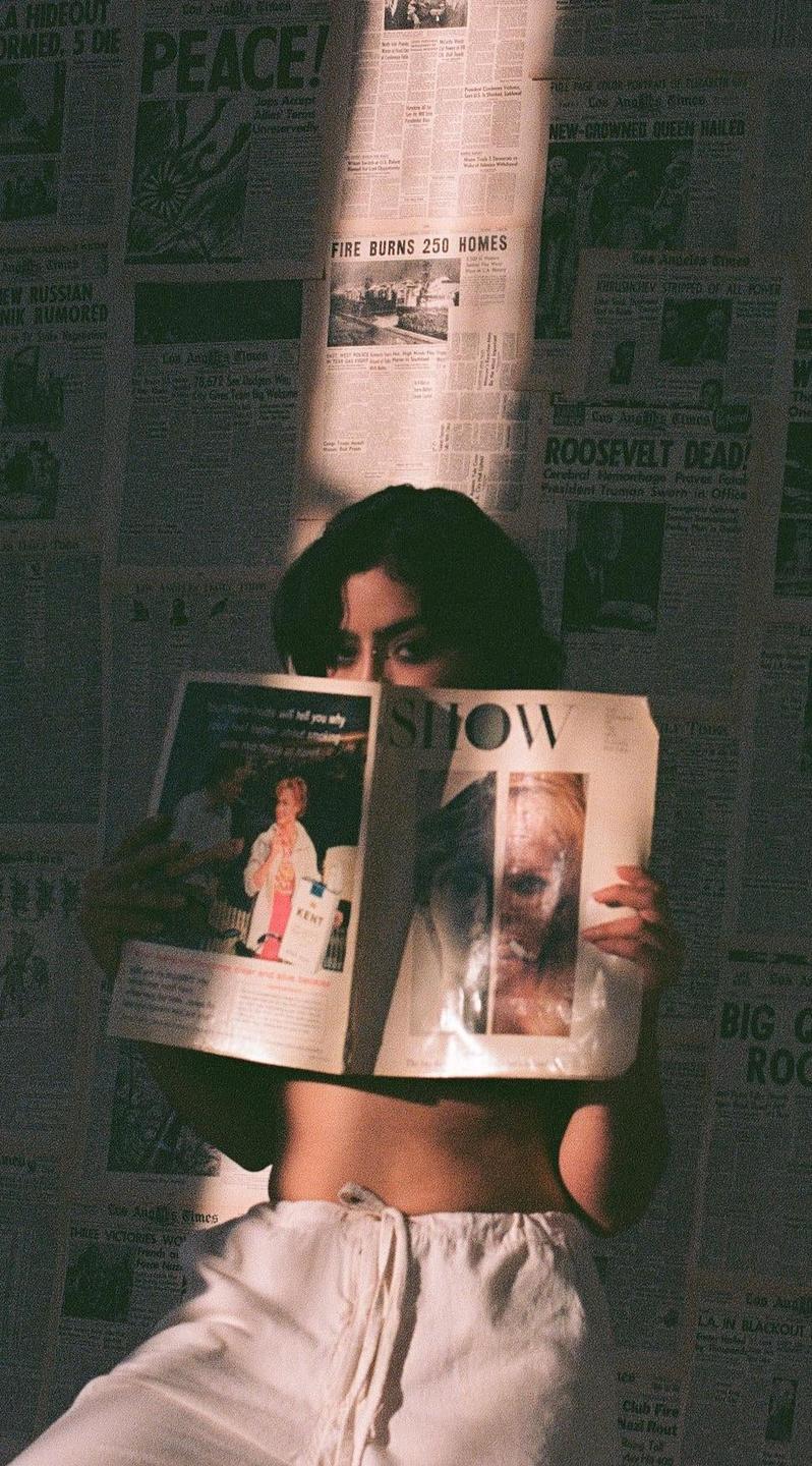 A woman holds a magazine in front of her face, with a wall covered in newspaper clippings behind her.