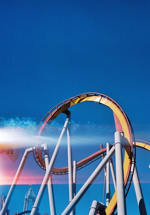 A yellow and red roller coaster track with white supports curves against a bright blue sky with a lens flare and light leak.
