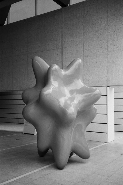 A large, smooth, abstract sculpture with multiple rounded protrusions stands on a tiled floor in front of a concrete wall.