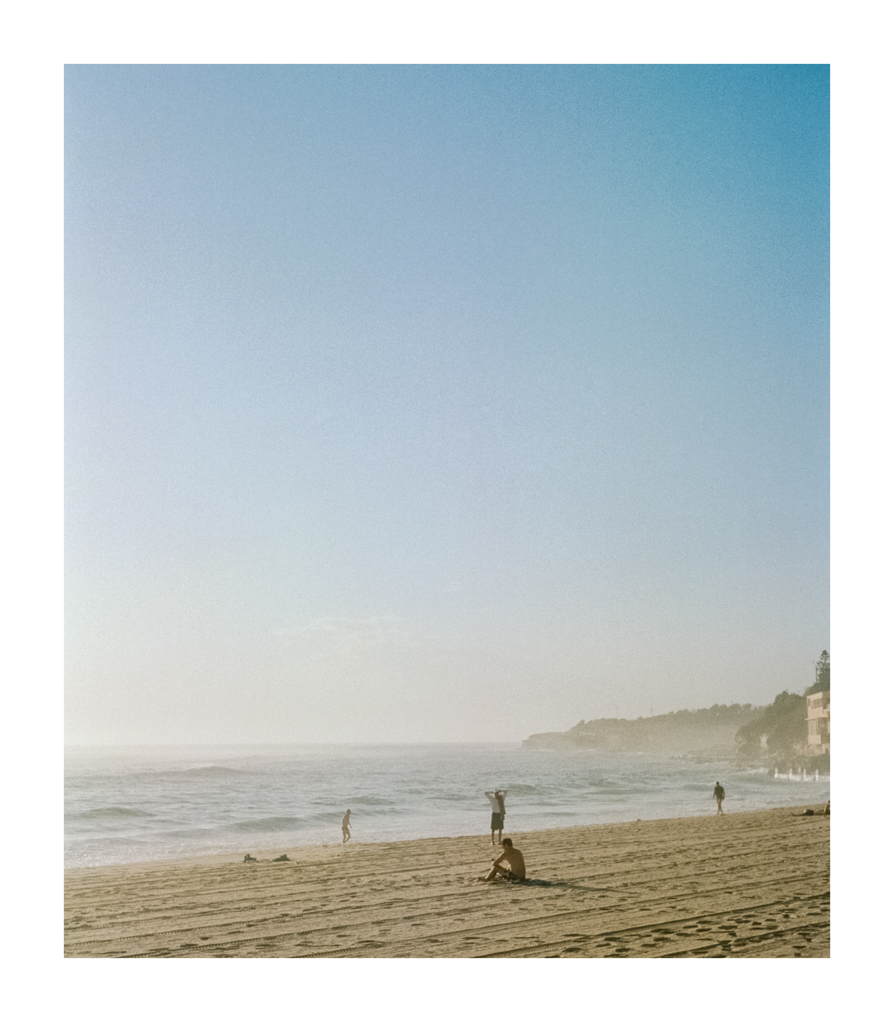 Several people are on a sandy beach with waves crashing on the shore and a hazy sky above.