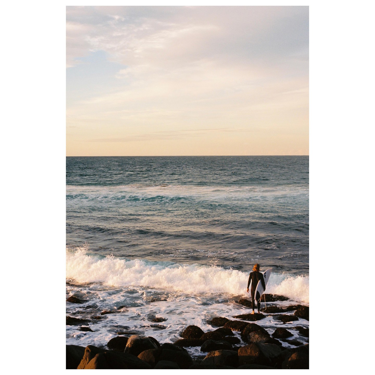 A surfer stands on rocks at the edge of the ocean, holding a surfboard and looking out at the waves.
