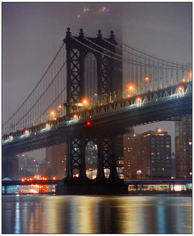 The Manhattan Bridge is illuminated at night, with its lights reflecting on the water below.