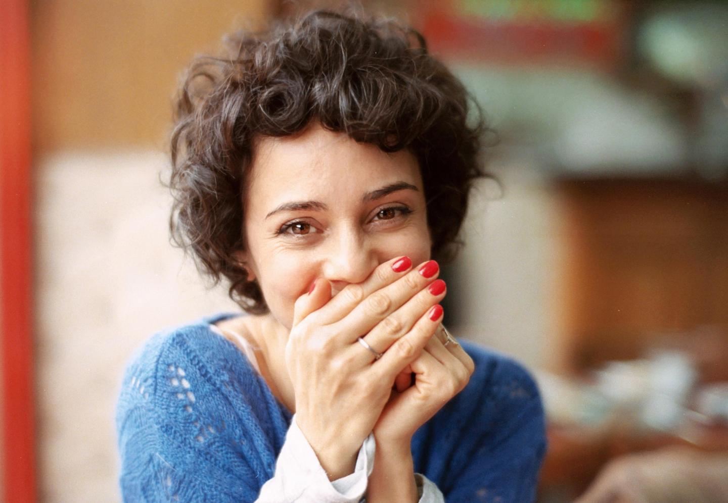 A woman with curly hair covers her mouth with her hands while smiling at the viewer.