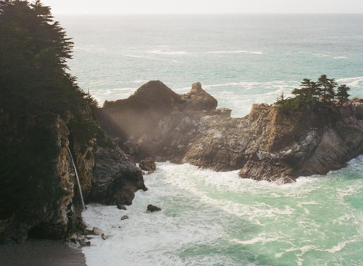 A waterfall cascades from a tree-covered cliff into the ocean, with waves crashing against rocky formations.