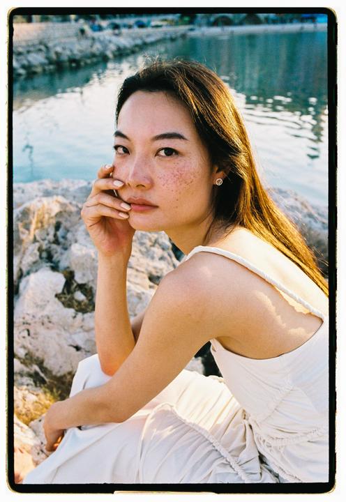 A woman with long hair and freckles sits on rocks by the water, looking towards the viewer.