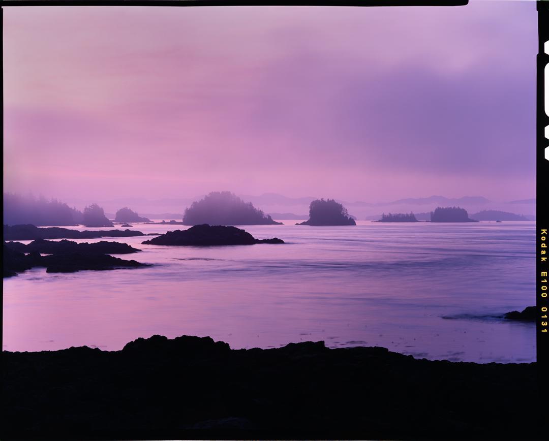 A purple sunrise illuminates a rocky coastline with small islands in the distance.
