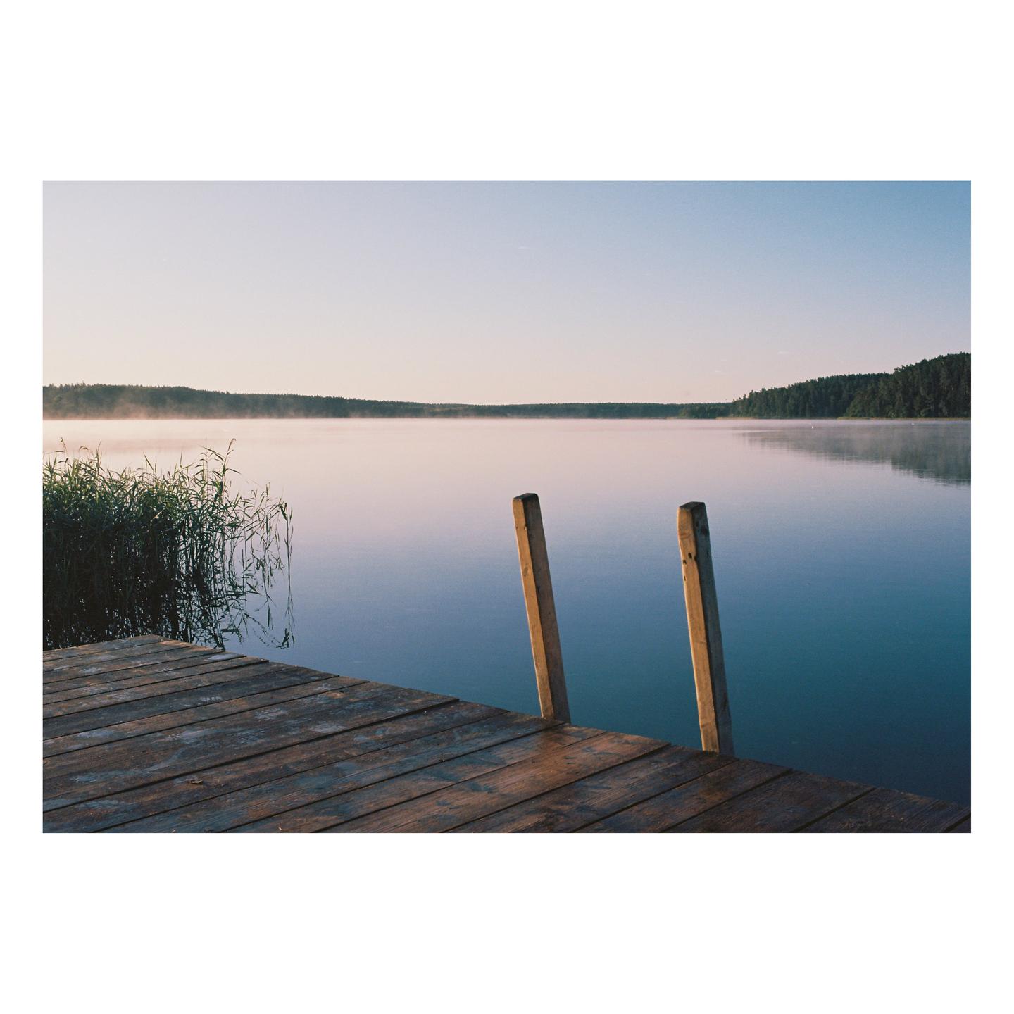 A wooden dock extends into a misty lake with a forest in the distance under a clear sky.