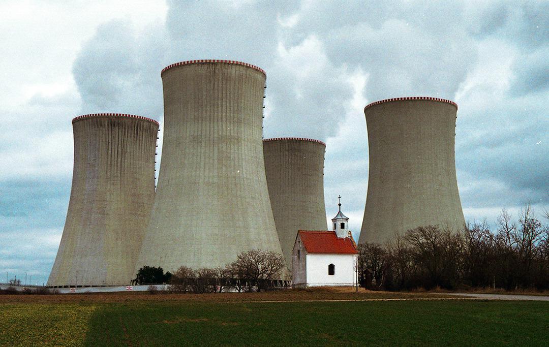Four cooling towers with smoke rising from them stand behind a small white chapel with a red roof.