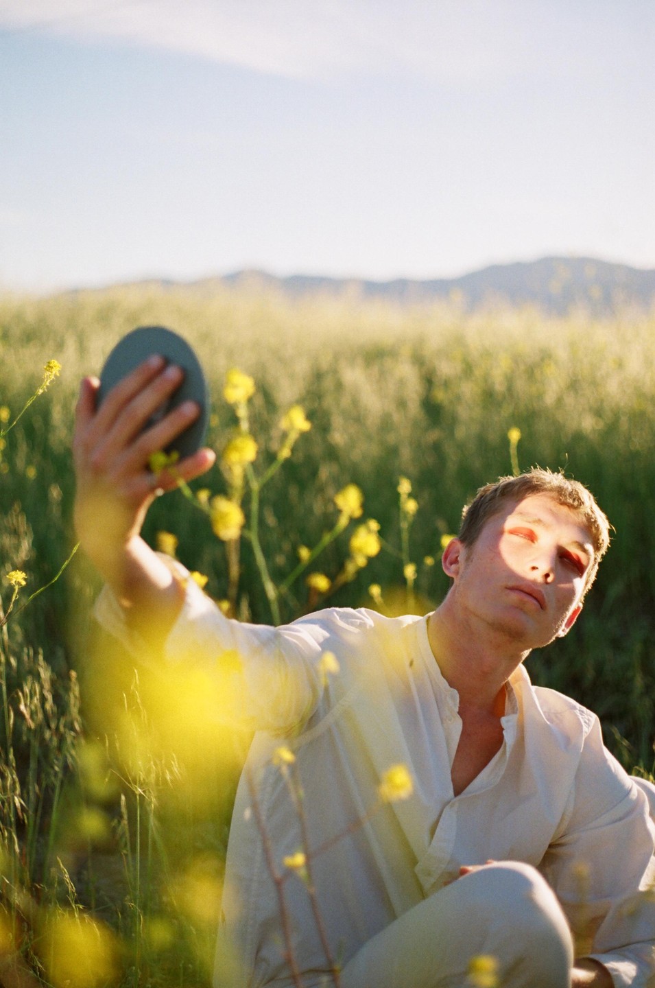 A man with red eyeshadow holds a mirror in a field of yellow flowers.