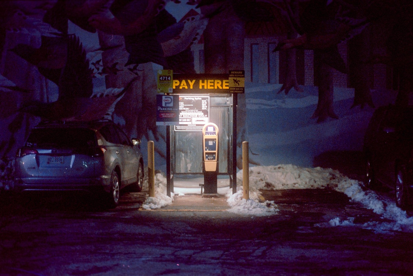 A parking payment machine stands illuminated at night next to a parked car, with snow on the ground and a mural on the wall.