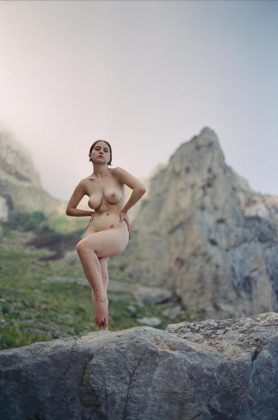 A nude woman stands on a rock with mountains in the background.