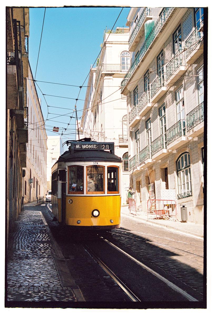 A yellow tram travels down a narrow, cobbled street lined with buildings under a clear sky.