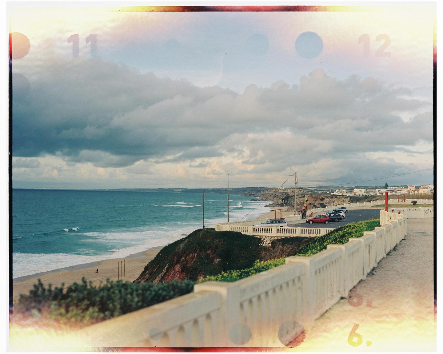 A coastal town with a beach and ocean is viewed from a high vantage point with a white fence in the foreground.
