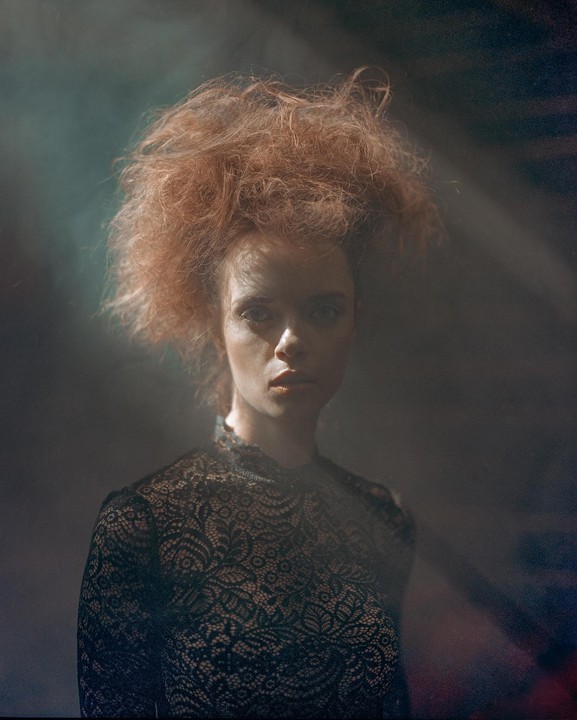 A woman with voluminous red hair and a black lace top is illuminated by a beam of light.