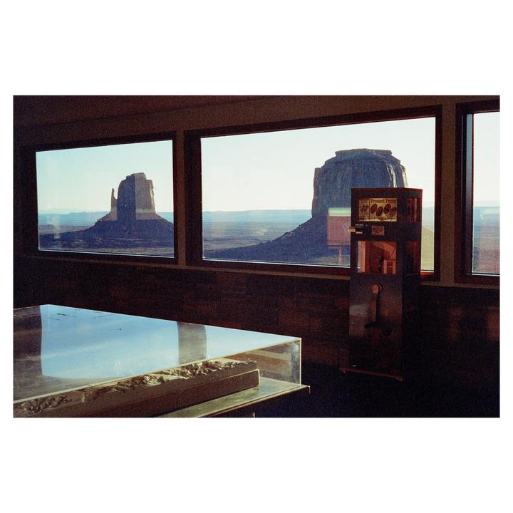 A view of Monument Valley through large windows shows two buttes and a vast desert landscape, with a display case and a penny press machine in the foreground.