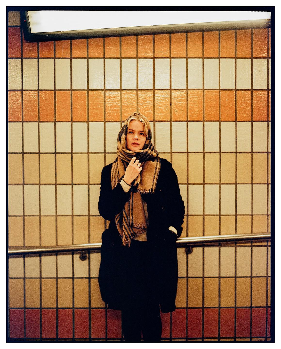 A woman wearing a plaid scarf and dark coat stands in front of a tiled wall.