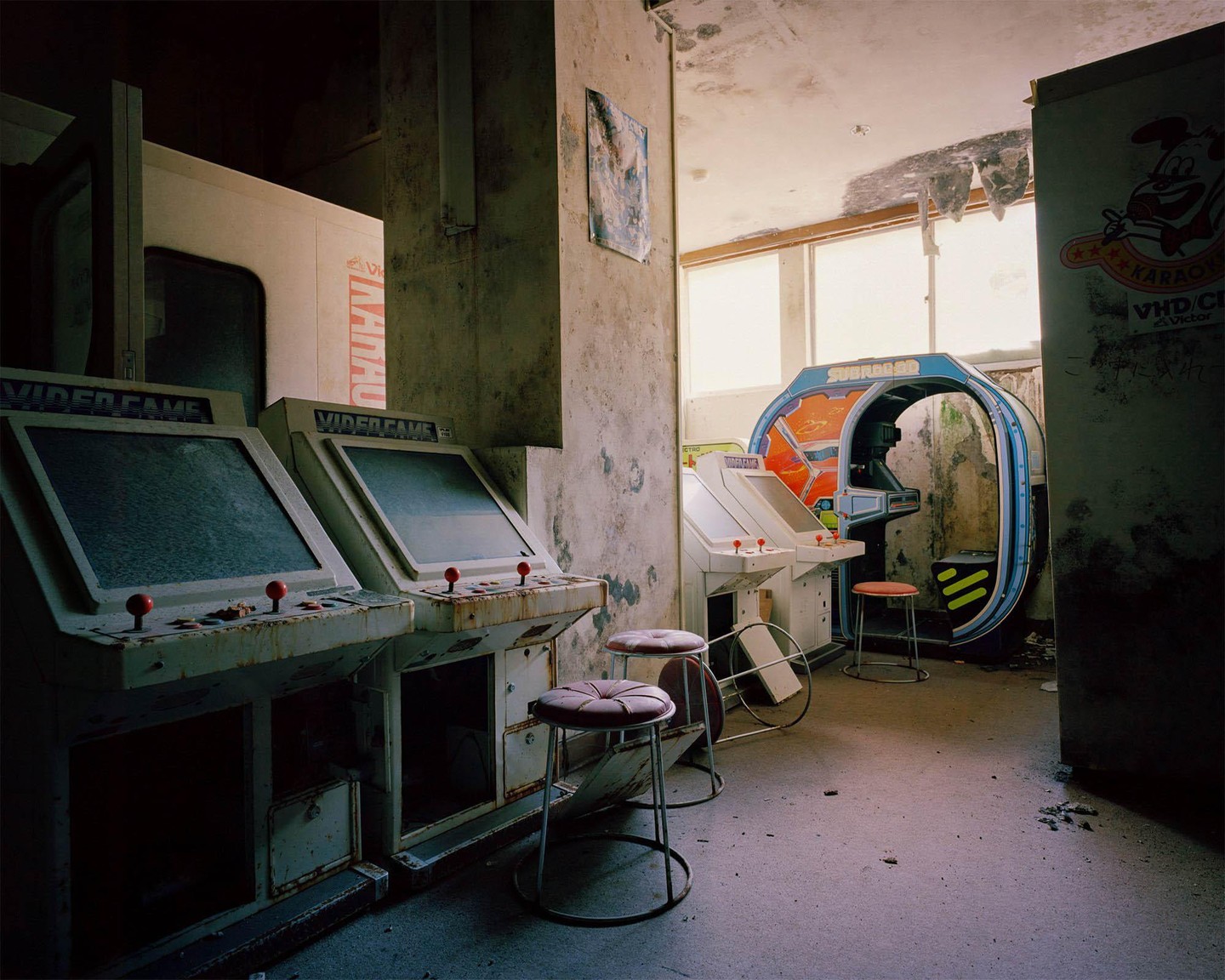 An abandoned arcade room features several dusty video game machines and stools, with peeling paint on the walls and ceiling.