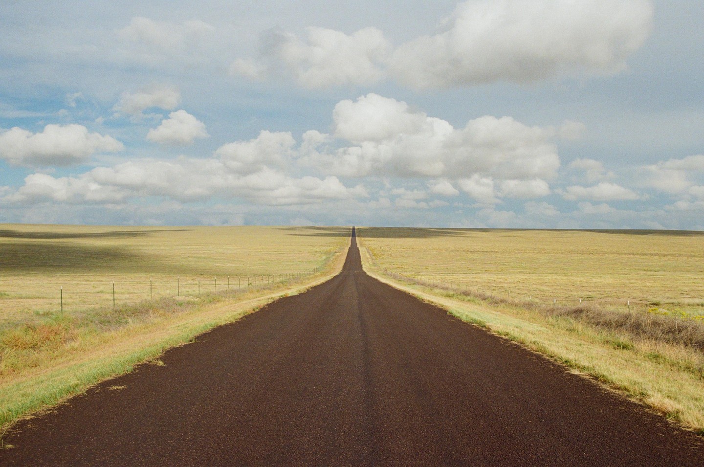 A long, straight road extends into the distance through a flat, grassy landscape under a cloudy sky.