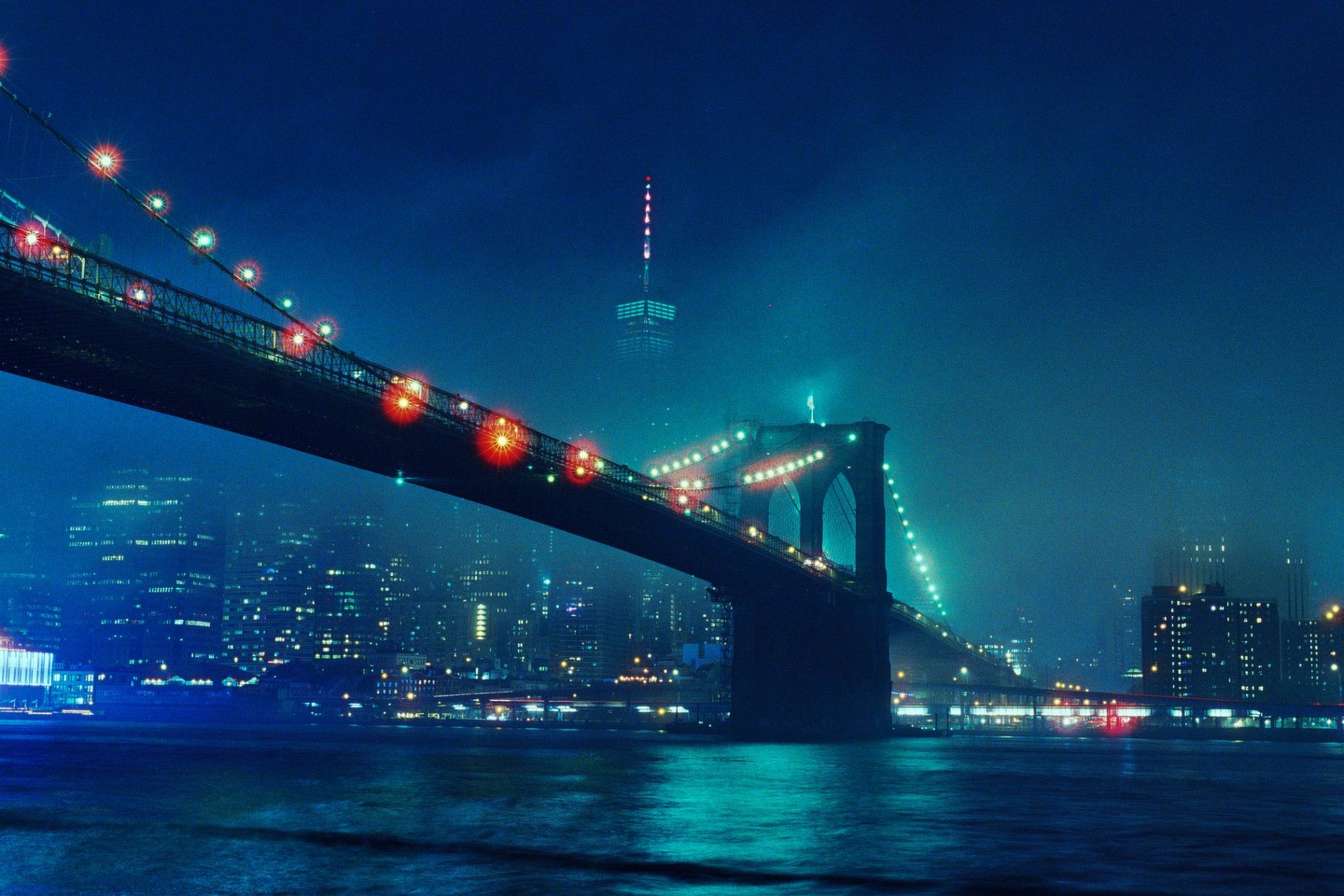 The Brooklyn Bridge is illuminated at night with a hazy city skyline in the background.