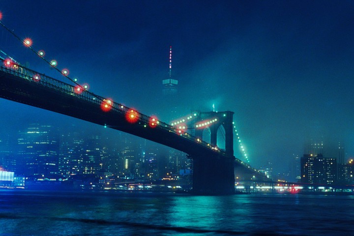 The Brooklyn Bridge is illuminated at night with a hazy city skyline in the background.