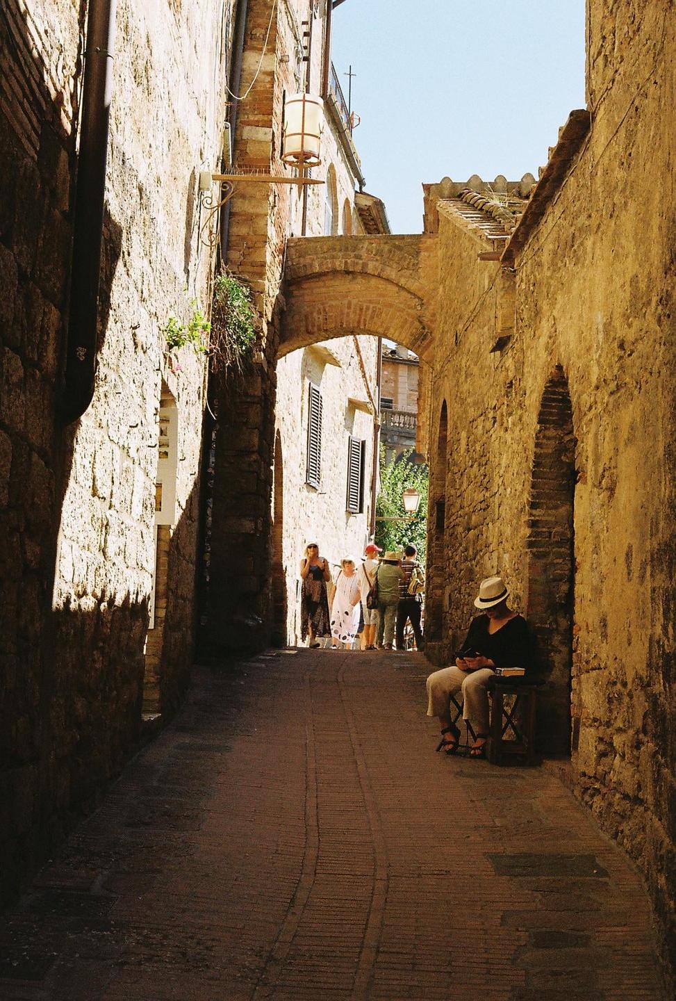 A man in a hat sits on a stool in a narrow, sunlit alleyway with a group of people in the distance.