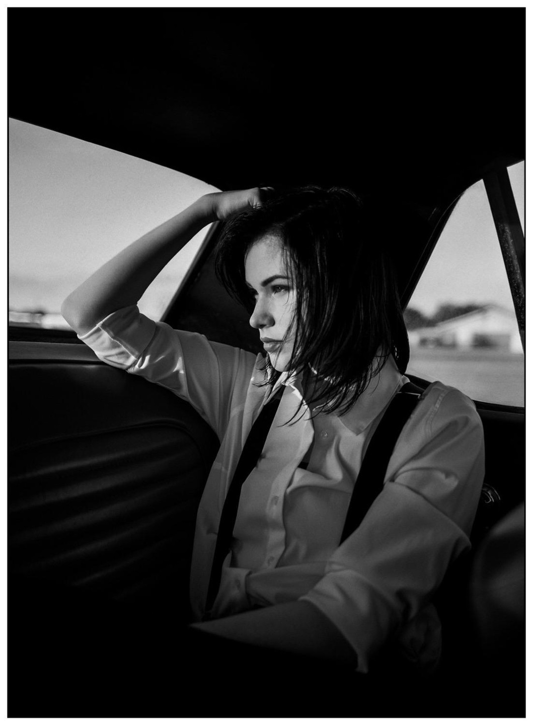 A woman with short hair and suspenders sits in the back of a car, looking out the window.