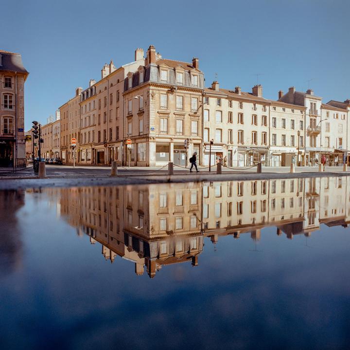 A person walks along a street with buildings reflected in a large puddle.