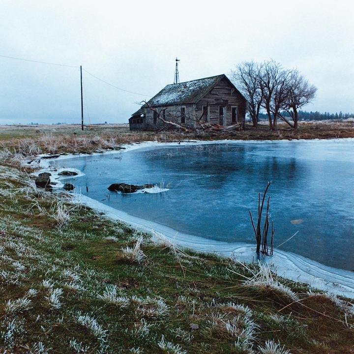 An abandoned wooden house with a windmill on its roof stands beside a partially frozen pond with frosted grass in the foreground.