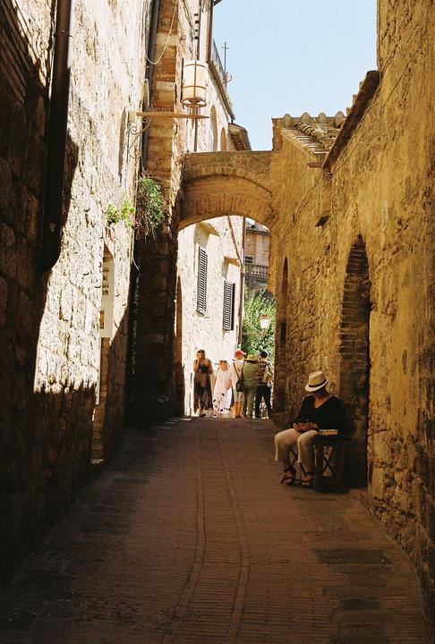 A man in a hat sits on a stool in a narrow, sunlit alleyway with a group of people in the distance.