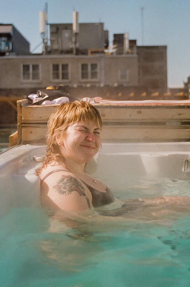 A woman with wet hair and a tattoo on her shoulder smiles while sitting in a hot tub on a rooftop.