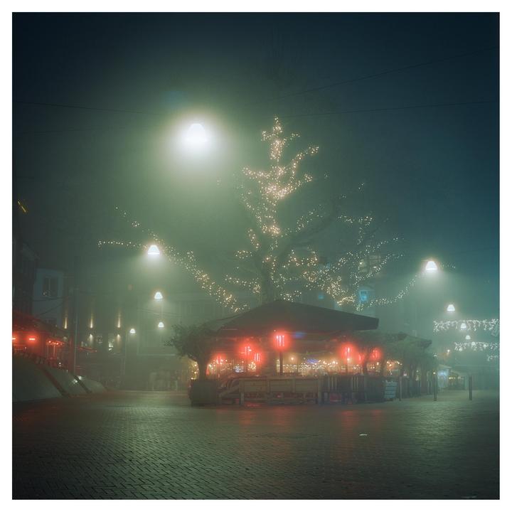 A street at night is illuminated by red and white lights, with a tree wrapped in lights in the background.