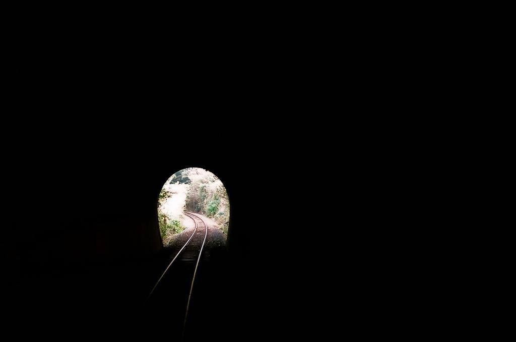 A view from inside a dark tunnel shows a bright opening with train tracks curving into a forest.