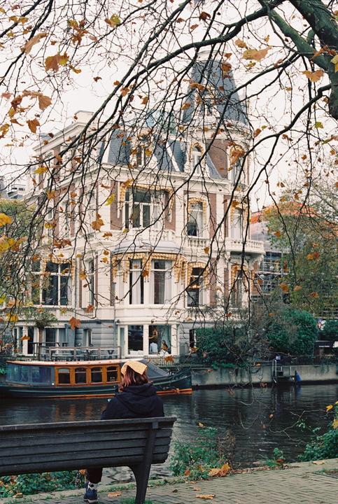 A person sits on a bench by a canal with a large building and a boat in the background.