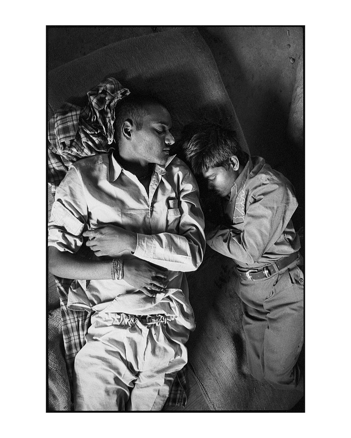 Two men sleep side by side on a textured surface, one with his arm across his chest and the other with his arm bent at his side.