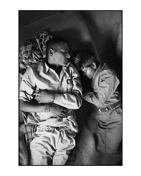 Two men sleep side by side on a textured surface, one with his arm across his chest and the other with his arm bent at his side.