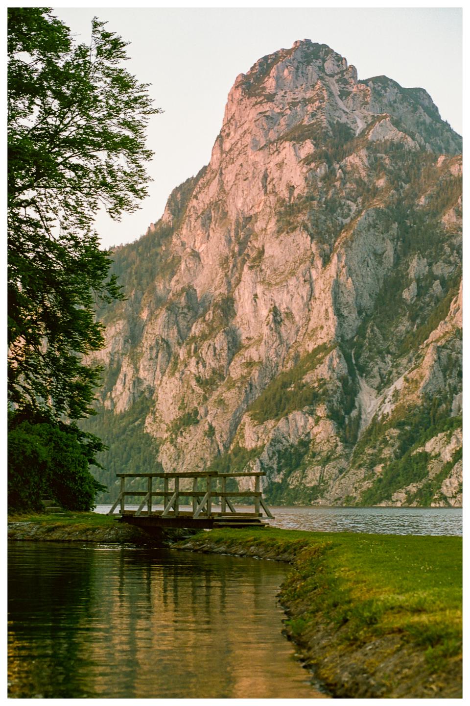 A wooden bridge crosses a narrow channel of water with a large mountain in the background.