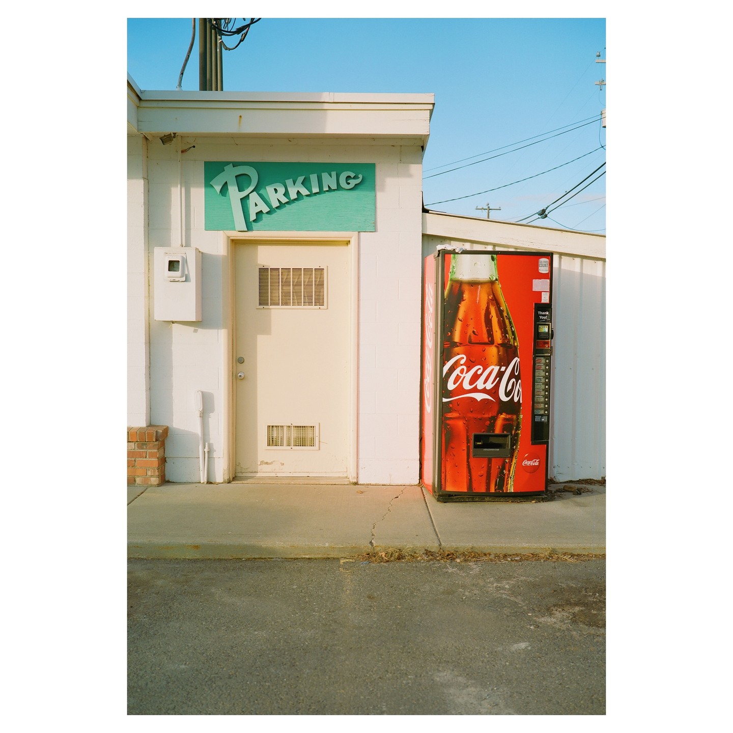 A red Coca-Cola vending machine stands next to a white building with a green "PARKING" sign above a door.