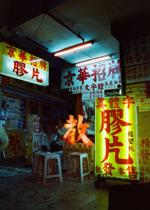 A man lights a cigarette in a shop surrounded by neon signs with Chinese characters.