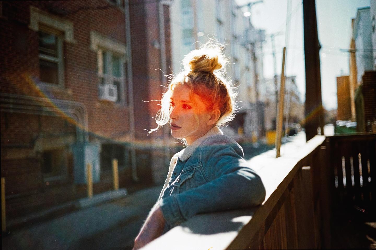 A woman with her hair in a bun leans on a wooden railing in an alleyway with brick buildings in the background.