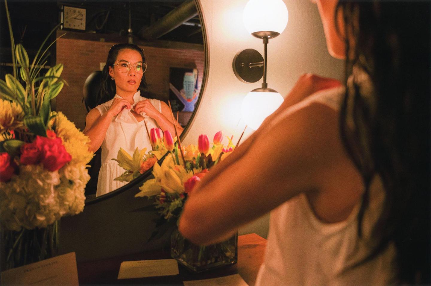 A woman in a white dress adjusts her collar while looking at her reflection in a mirror, with bouquets of flowers on either side.