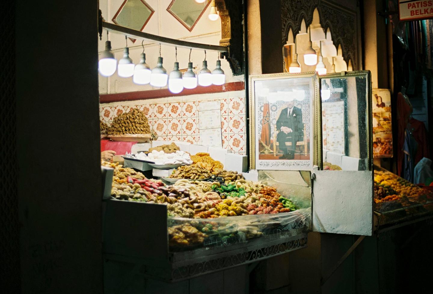 A brightly lit stall in a market displays a variety of colorful pastries and a framed portrait of a man.