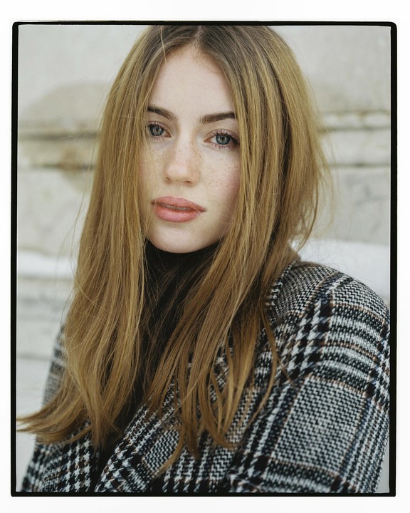 A woman with long hair and a plaid coat looks directly at the viewer.