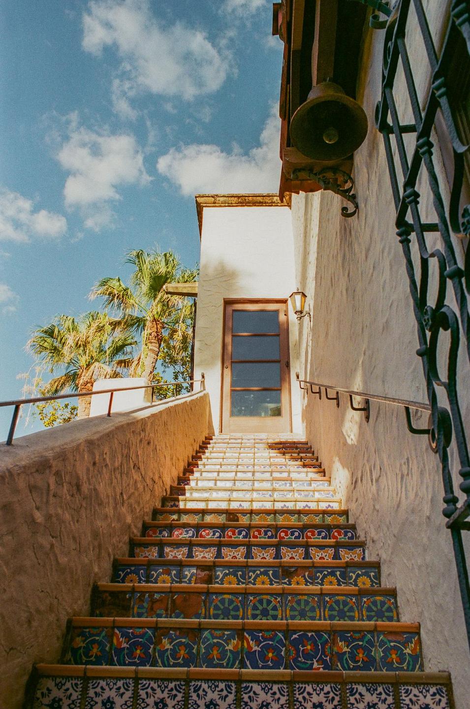 A long outdoor staircase with colorful patterned tiles leads up to a wooden door on a building with palm trees and a blue sky in the background.